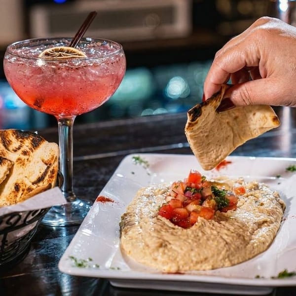 A hand dips pita bread into hummus topped with tomatoes, next to a pink cocktail.