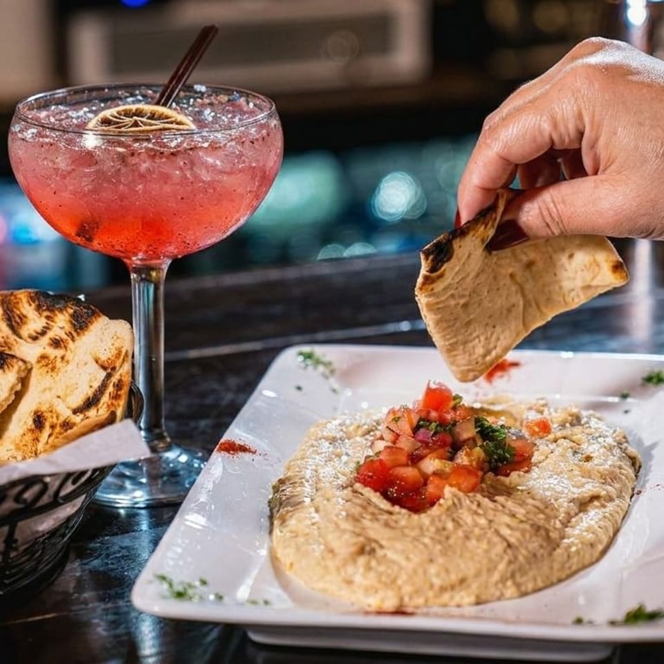 A hand dips pita bread into hummus topped with tomatoes, next to a pink cocktail.