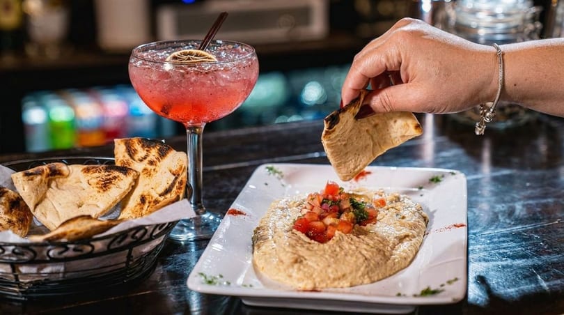 A hand dips pita bread into hummus topped with tomatoes, next to a pink cocktail.