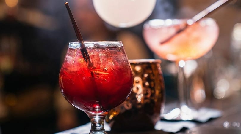 Three cocktails on a bar, a red drink with a straw in the foreground.