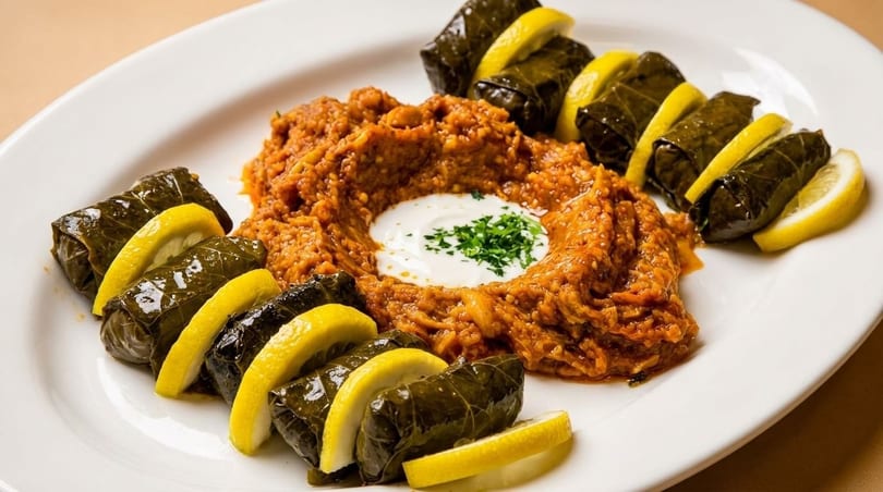 Grape leaves with lemon slices surround a mound of red lentil kofte topped with yogurt and parsley.