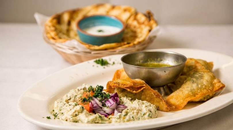 A plate of fried dumplings, a creamy dip with red onions, and a basket of flatbread with a dip.