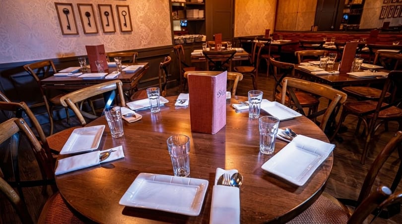 Cozy restaurant interior with round wooden tables set with white plates, glasses, and menus.