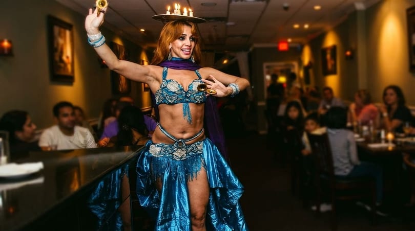 A belly dancer in a blue costume with candles on her head performs for restaurant patrons.